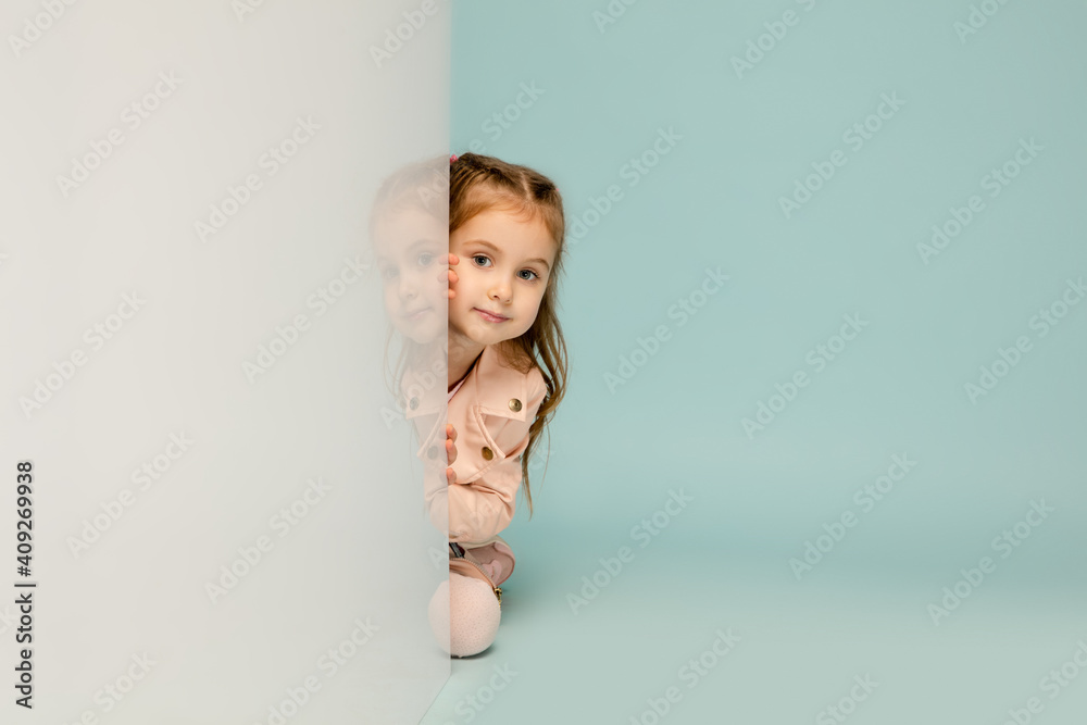 Lovely. Happy kid, girl isolated on blue studio background. Looks happy, cheerful. Copyspace for ad. Childhood, education, emotions, facial expression concept. Peeking out from behind the wall. Lovely. Happy kid, girl isolated on blue studio background. Looks happy, cheerful. Copyspace for ad. Childhood, education, emotions, facial expression concept. Peeking out from behind the wall.