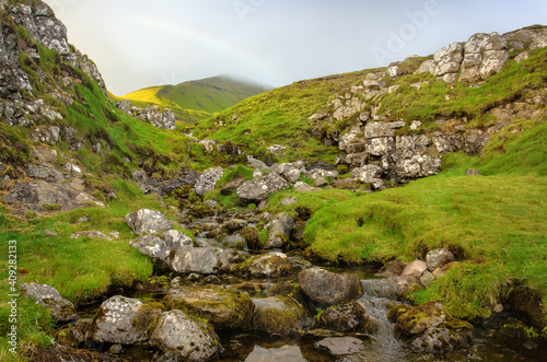 The landscape of the Faroe Islands - a small river in a rocky slope overlooking the top of a hill with a rainbow in the sky.