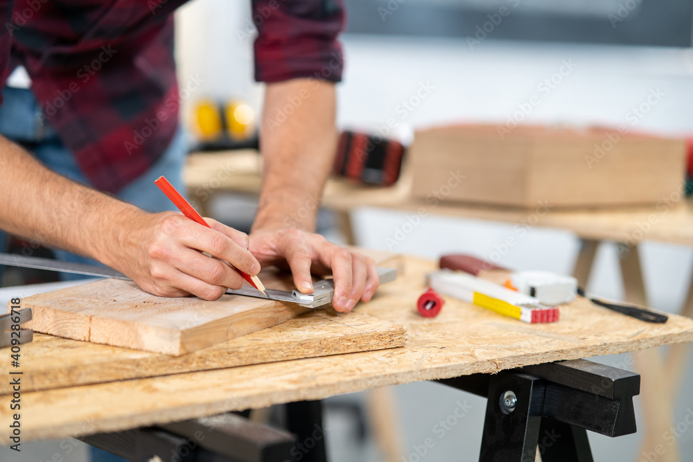 Close-up of a hardworking professional carpenter holding a angular ruler and pencil while measuring a board in a carpentry workshop. A bearded DIY enthusiast measures wood. There are a locksmith table Close-up of a hardworking professional carpenter holding a angular ruler and pencil while measuring a board in a carpentry workshop. A bearded DIY enthusiast measures wood. There are a locksmith table