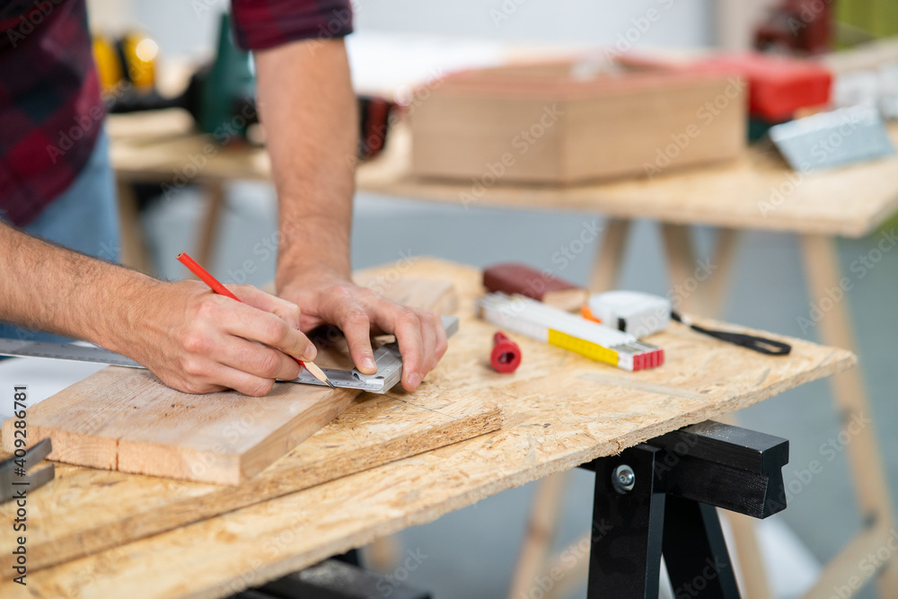 Close-up of a hardworking professional carpenter holding a angular ruler and pencil while measuring a board in a carpentry workshop. A bearded DIY enthusiast measures wood. There are a locksmith table Close-up of a hardworking professional carpenter holding a angular ruler and pencil while measuring a board in a carpentry workshop. A bearded DIY enthusiast measures wood. There are a locksmith table