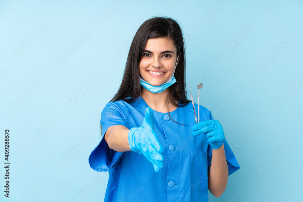Young woman dentist holding tools over isolated blue background handshaking after good deal Young woman dentist holding tools over isolated blue background handshaking after good deal