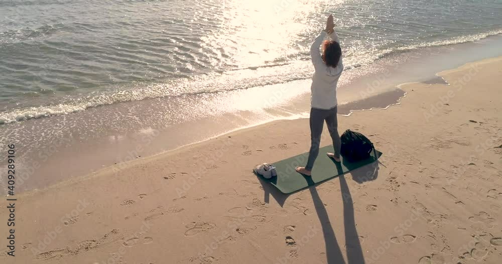 Mujer mediana edad haciendo yoga delante del mar y el sol y dron orbitando
