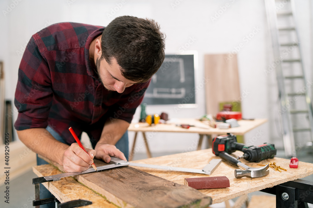 Close-up of a hardworking professional carpenter holding a angular ruler and pencil while measuring a board in a carpentry workshop. A bearded DIY enthusiast measures wood. There are a locksmith table Close-up of a hardworking professional carpenter holding a angular ruler and pencil while measuring a board in a carpentry workshop. A bearded DIY enthusiast measures wood. There are a locksmith table