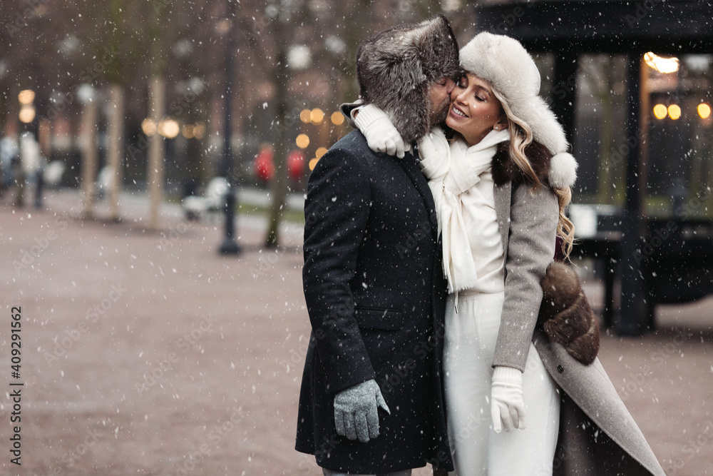 Fashion couple of lovers embracing, kissing, standing on winter street in snowy weather, man and woman wear fur hat and warm wool coat. St. Valentines Day, Christmas holidays concept. 