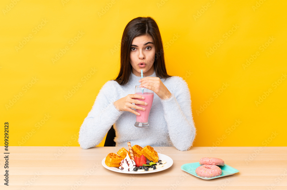 Young woman eating waffles and milkshake in a table over isolated yellow background