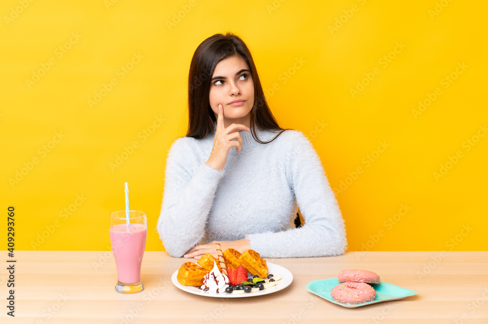 Young woman eating waffles and milkshake in a table over isolated yellow background thinking an idea Young woman eating waffles and milkshake in a table over isolated yellow background thinking an idea