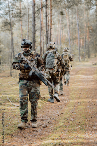 Squad of Four Fully Equipped Soldiers in Camouflage on a Reconnaissance Military Mission, Aiming Rifles. They're Moving in Formation Through Dense Pine Forest.