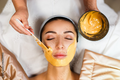 Cosmetic jelly mask, facial skincare. Cosmetologist applies yellow gold alginate mask with spatula on face of a woman. Facial skin treatment in the beauty salon. Top View above