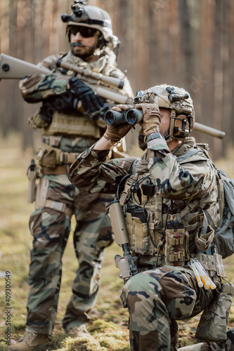Portrait of a bearded middle-aged soldier in a Woodland military uniform and a helmet with headphones on his head, holding a rifle and looking around through the thick pine woods.