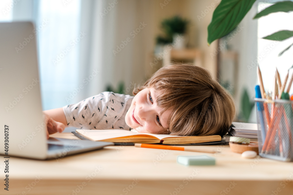 Distance learning online education. Caucasian smile kid boy studying at home, tired and lying on laptop and doing school homework. Thinking child siting at table with notebook. Back to school. Distance learning online education. Caucasian smile kid boy studying at home, tired and lying on laptop and doing school homework. Thinking child siting at table with notebook. Back to school.