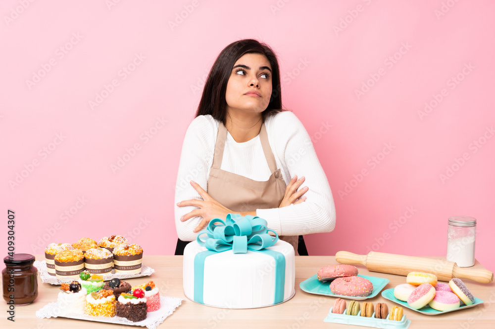Pastry chef with a big cake in a table over isolated pink background making doubts gesture while lifting the shoulders Pastry chef with a big cake in a table over isolated pink background making doubts gesture while lifting the shoulders