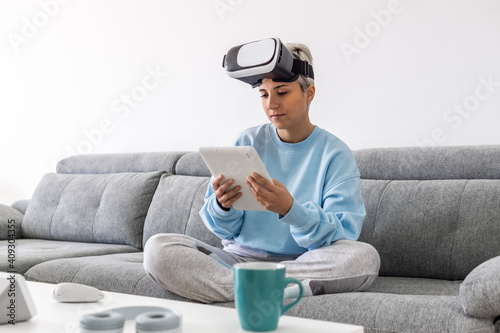 Beautiful girl on the couch at home wearing virtual reality glasses and browsing the internet with a tablet