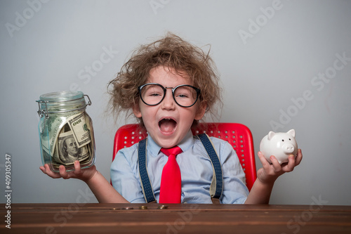 Kid and money. Funny excited child plays with money. Education for little businessman how to be rich when grow up. Student in glasses and tie with piggy bank and jar full of money