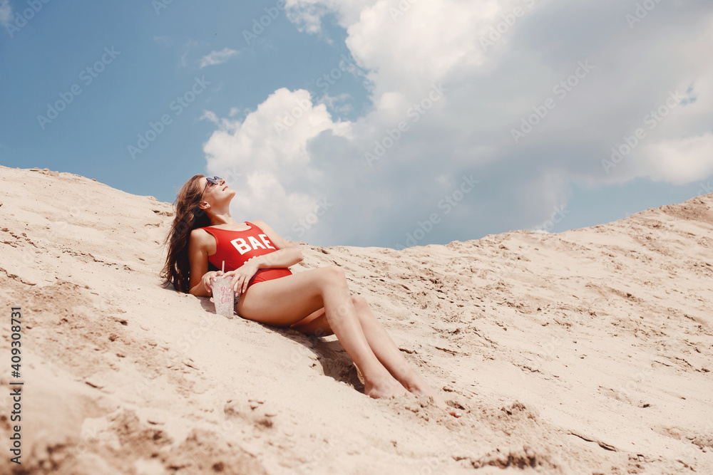 Relaxing on beach, young woman lies on sand in red swimsuit with glass of cocktail and sunglasses Relaxing on beach, young woman lies on sand in red swimsuit with glass of cocktail and sunglasses
