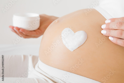 Girl sitting in bed. Young adult woman hands holding opened white cream jar. Heart shape created from cream on naked big belly. Care about perfect, soft and smooth skin in pregnancy time. Close up.