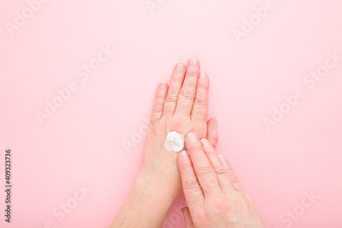 Old mature woman hands using anti aging moisturizing white cream on light pink table background. Pastel color. Care about clean and soft body skin. Daily beauty product. Closeup. Top down view.