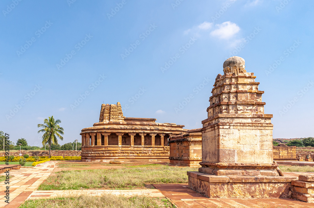 Aihole, Karnataka, India - November 7, 2013: Brown stone Durga Gudi or Temple under blue cloudscape. Small shrine in front and some green foliage and grass.
