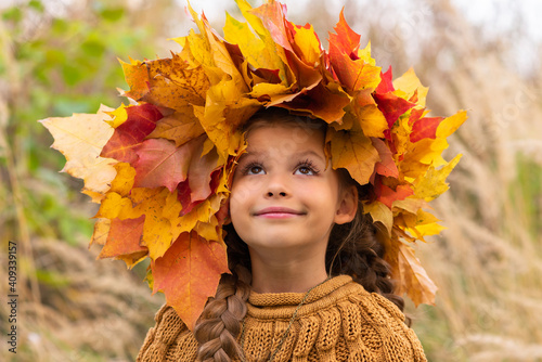 A little girl with a wreath of maple leaves on her head in a warm sweater on an autumn day.