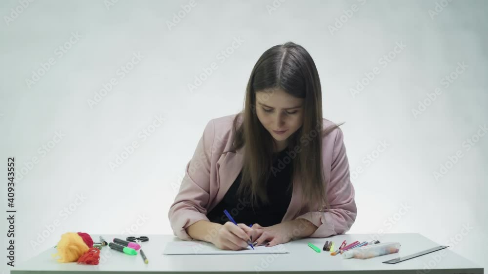 Attractive young woman uses markers to draw picture on on a white paper at the table in a white studio Attractive young woman uses markers to draw picture on on a white paper at the table in a white studio