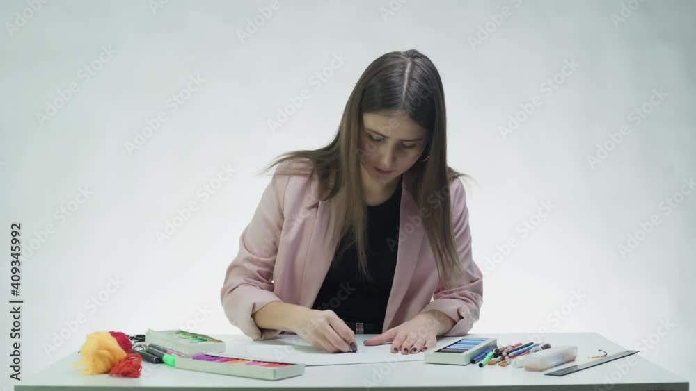 Attractive young woman uses pastel to draw something on on a white paper at the table in a white studio Attractive young woman uses pastel to draw something on on a white paper at the table in a white studio