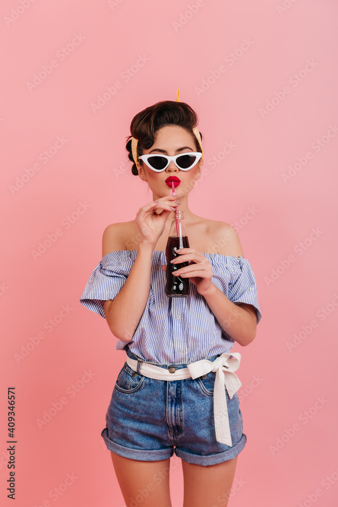 Shapely girl in denim shorts and striped blouse drinking beverage. Studio shot of pinup lady in sunglasses.