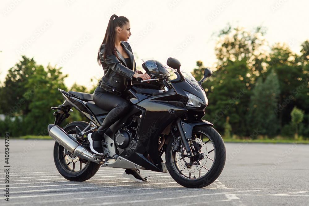 Stylish beautiful girl in black leather jacket and pants on outdoors parking sits on sports motorcycle and holds protective helmet. Stylish beautiful girl in black leather jacket and pants on outdoors parking sits on sports motorcycle and holds protective helmet.