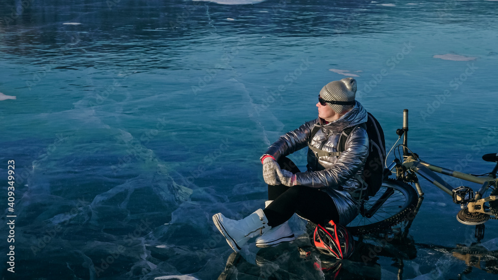 Woman sitting near her bike on ice. The girl cyclist stopped to rest. She sits on the wheel and enjoys the beautiful view of the sunset. The traveler is ride a cycle. Female is riding bike on the ice. Woman sitting near her bike on ice. The girl cyclist stopped to rest. She sits on the wheel and enjoys the beautiful view of the sunset. The traveler is ride a cycle. Female is riding bike on the ice.