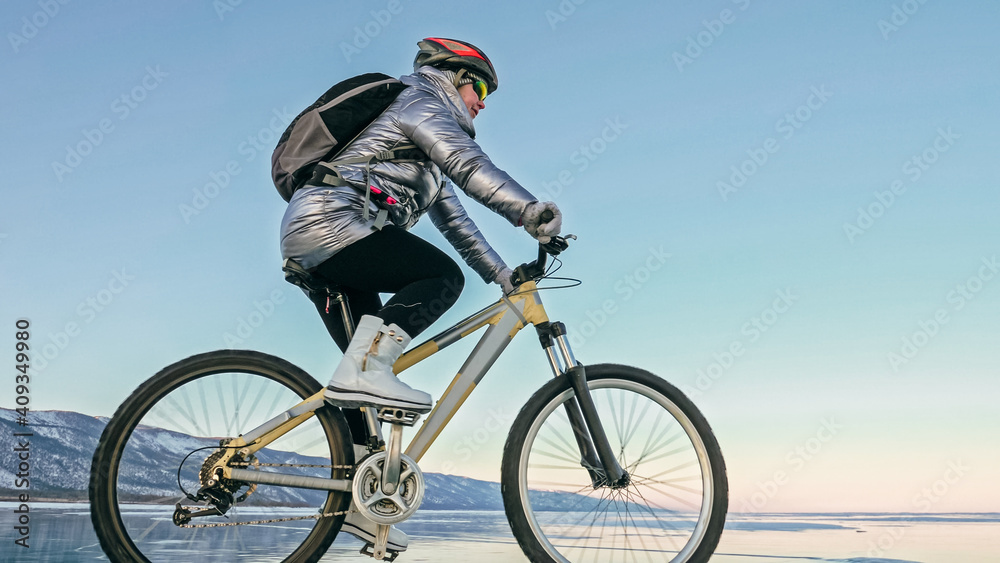 Obraz premium Woman is riding bicycle on the ice. Girl is dressed in a silvery down jacket, cycling backpack and helmet. Ice of the frozen Lake Baikal. Tires on bike are covered with spikes. Traveler is ride cycle.