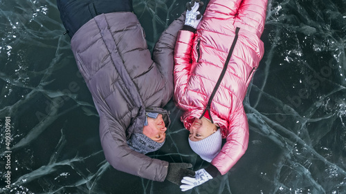 Couple has fun winter walk against background of ice of frozen lake. Lovers lie on clear ice with cracks have fun kiss and hug. View from above. Happy people on snow covered ice. Honeymoon love story.