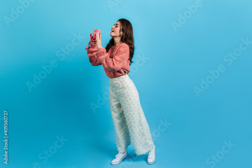 Full-length shot of charming brunette in white pants and pink sweater. Girl with interest takes picture on Instax