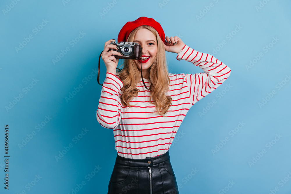 Carefree french girl holding camera on blue background. Studio shot of blonde woman in beret taking photos. Carefree french girl holding camera on blue background. Studio shot of blonde woman in beret taking photos.
