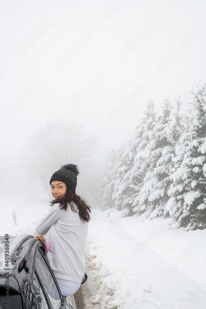 Junge Frau sitzt auf dem Auto in der Winterlandschaft. Sie ist weiß mit einer schwarzen Mütze angezogen. Im Hintergrund verschneite Landschaft. Sie schaut in die Kamera. Junge Frau sitzt auf dem Auto in der Winterlandschaft. Sie ist weiß mit einer schwarzen Mütze angezogen. Im Hintergrund verschneite Landschaft. Sie schaut in die Kamera.