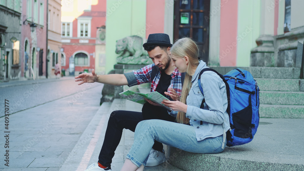 Happy tourist couple sitting on street stairs, checking city map and discussing. In the background there is beautiful old town view. Happy tourist couple sitting on street stairs, checking city map and discussing. In the background there is beautiful old town view.