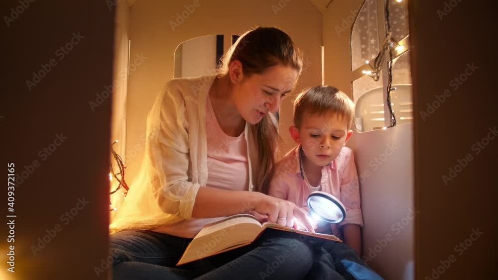 Portrait of smiling young mother with little boy reading book at night while playing in toy house. Concept of child education and family having time together at night Portrait of smiling young mother with little boy reading book at night while playing in toy house. Concept of child education and family having time together at night