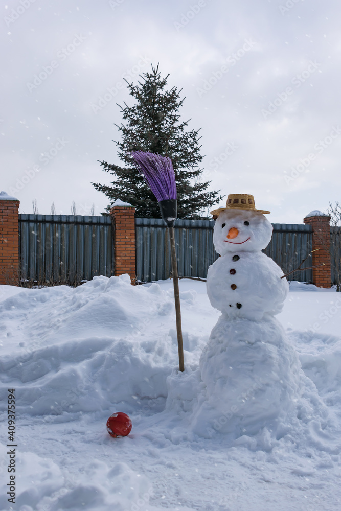 Obraz premium A snowman in a straw hat and with a carrot nose in the winter garden, holding a broom in his 