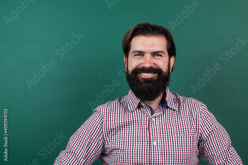 cheerful mature teacher on background of blackboard. brutal bearded man wear casual checkered shirt and smiling. express positive human emotions. caucasian hipster on green background. copy space