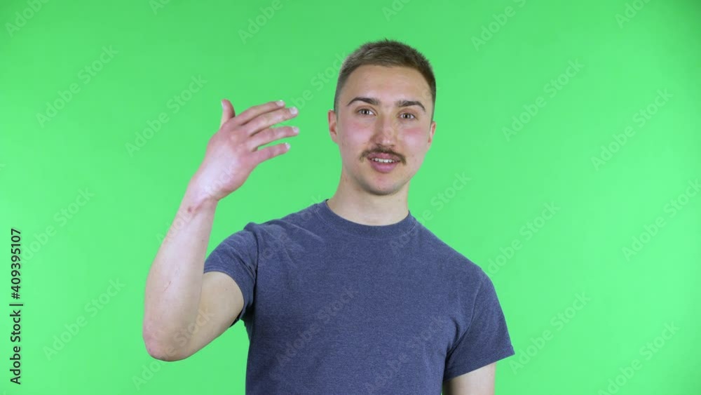 Portrait of a young man looking at the camera waving hello and making gesture come here. Cute male with a mustache in a blue t-shirt posing on a green screen in the studio. Close up. Portrait of a young man looking at the camera waving hello and making gesture come here. Cute male with a mustache in a blue t-shirt posing on a green screen in the studio. Close up.