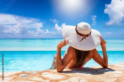 A woman with white hat sits by the pool and enjoys the view to the tropical, turquoise sea during her summer holiday © moofushi