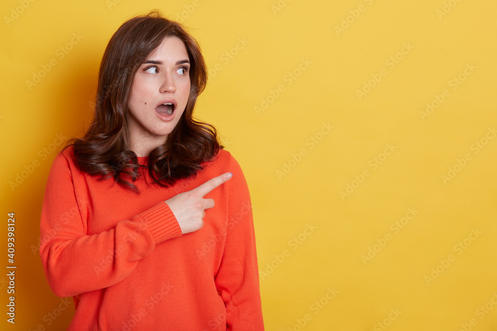 Portrait of shocked caucasian female with pleasant appearance indicates aside at blank yellow wall, has surprised and astonished expression, demonstrates something amazing.