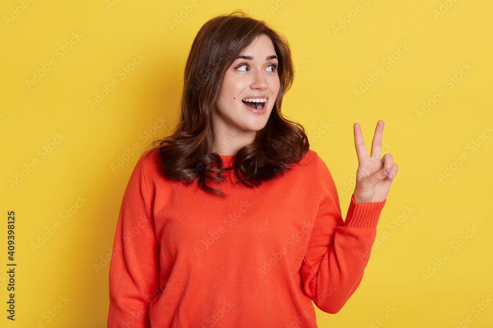 Portrait of joyful smiling pretty girl wearing orange jumper, showing v sign with fingers and looking aside with excited facial expression, shows peace or victory gesture, rejoicing success. Portrait of joyful smiling pretty girl wearing orange jumper, showing v sign with fingers and looking aside with excited facial expression, shows peace or victory gesture, rejoicing success.