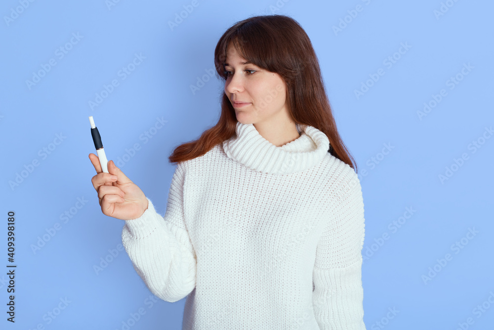 Young dark haired lady vaping, holding electric cigarette in hands and looking at it, woman wearing white casual clothing posing indoor isolated over blue background. Young dark haired lady vaping, holding electric cigarette in hands and looking at it, woman wearing white casual clothing posing indoor isolated over blue background.