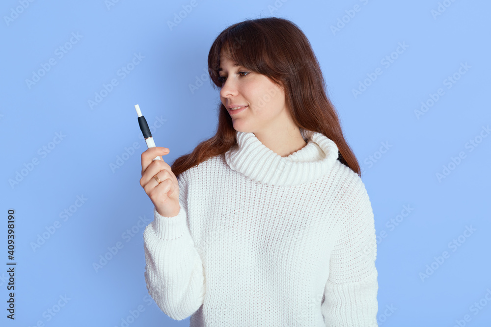 Young attractive female with pleasant appearance, lady wearing white sweater standing against blue wall and looking aside, girl with dark hair decides smoke or not.