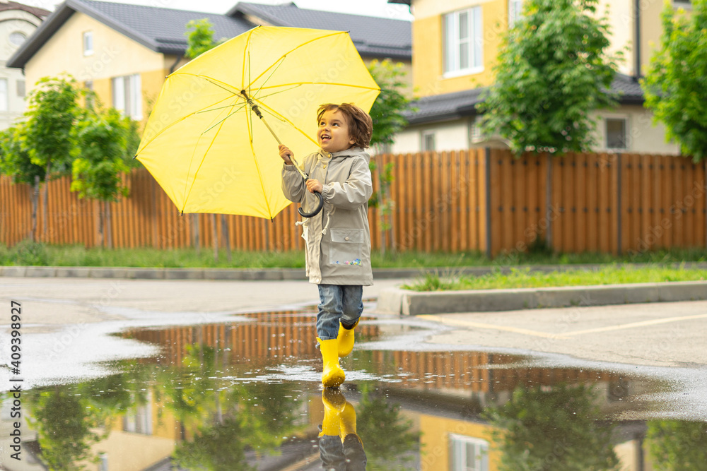 Baby gay boy runs through puddles after rain in the street with yellow umbrella and boots