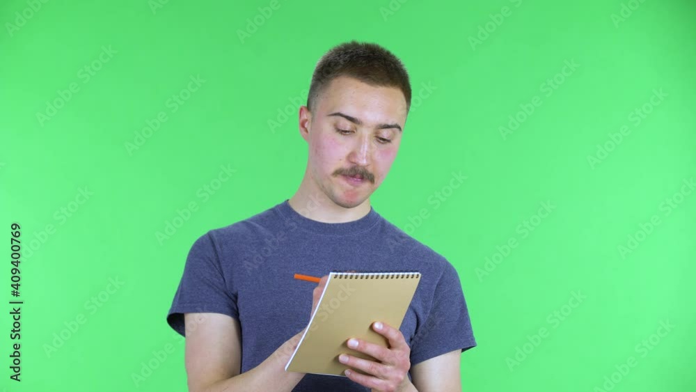 Portrait of a young man is thinking, then happy writing with pencil in notebook. Cute male with a mustache in a blue t-shirt posing on a green screen in the studio. Close up. Portrait of a young man is thinking, then happy writing with pencil in notebook. Cute male with a mustache in a blue t-shirt posing on a green screen in the studio. Close up.