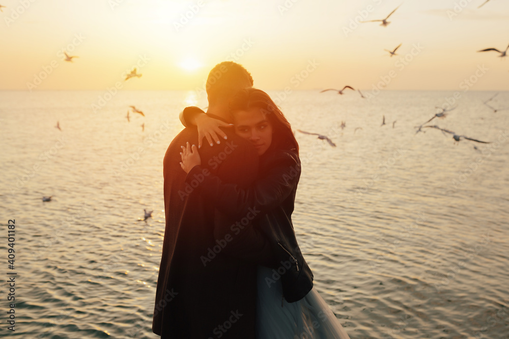 Silhouette of a young couple in love hugging on the sea beach at sunset with flying seagulls on the background.