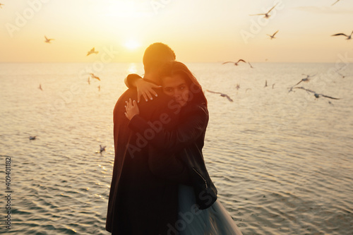 Obraz na płótnie Silhouette of a young couple in love hugging on the sea beach at sunset with flying seagulls on the background. w salonie
