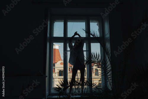 Silhouette of slim girl with hands up standing on the windowsill and dancing at home.