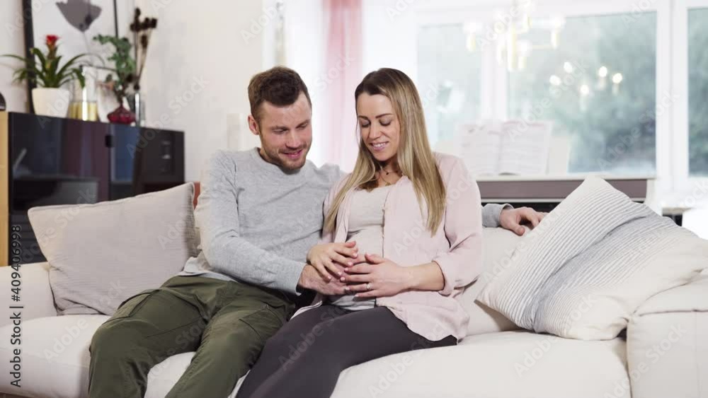 Young pregnant happy couple sitting on couch husband stroking baby belly of blond wife shot in 4k Young pregnant happy couple sitting on couch husband stroking baby belly of blond wife shot in 4k
