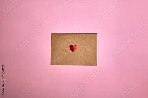 A red heart lies on a brown envelope for Valentine's Day. Pink backdrop with envelope and red heart. Love letter or message concept. Flat-lay, top view. Copy space for your text.