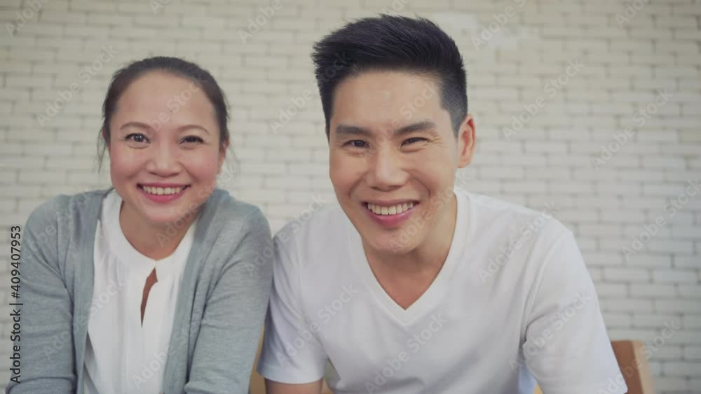 Young asian couple smiling and waving while on video call with overseas family using laptop sitting on sofa in the living room at home. Filling their home with love. Young couple working at home. Young asian couple smiling and waving while on video call with overseas family using laptop sitting on sofa in the living room at home. Filling their home with love. Young couple working at home.